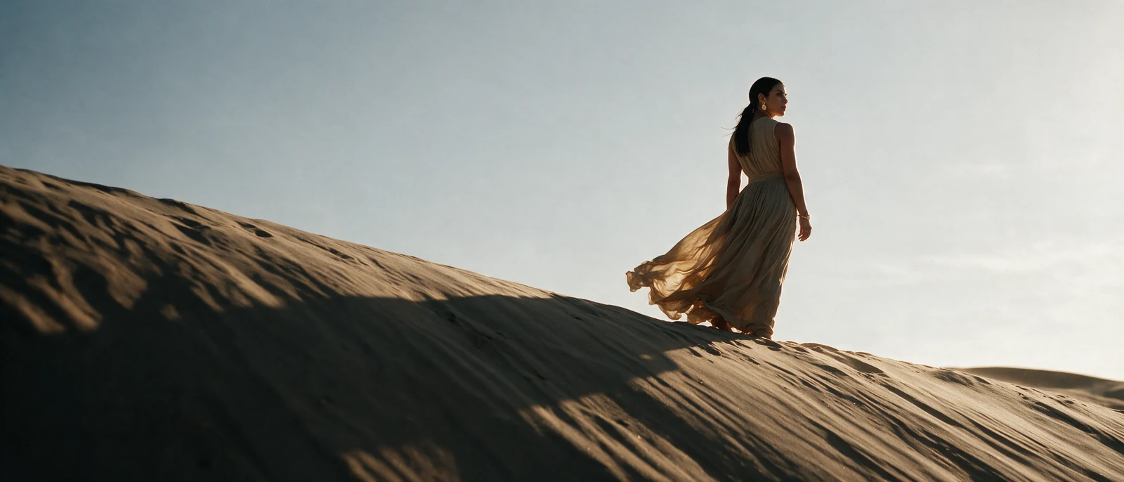 A woman in a pale, wind-blown dress stands on the crest of a dune, seen from behind, looking out over the sand.