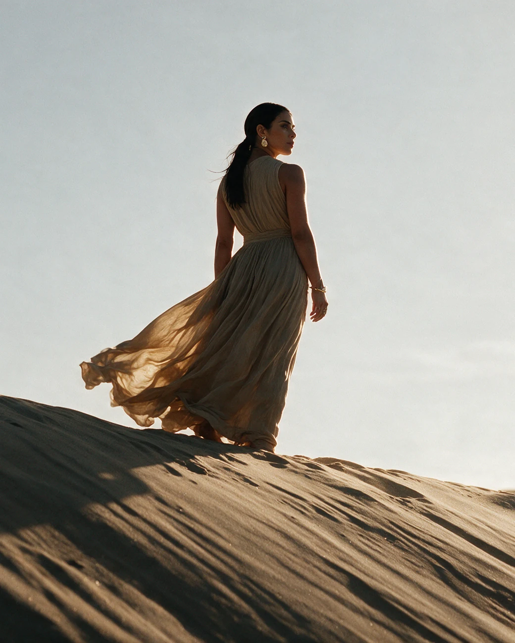 She stands on a dune crest, seen from behind.
