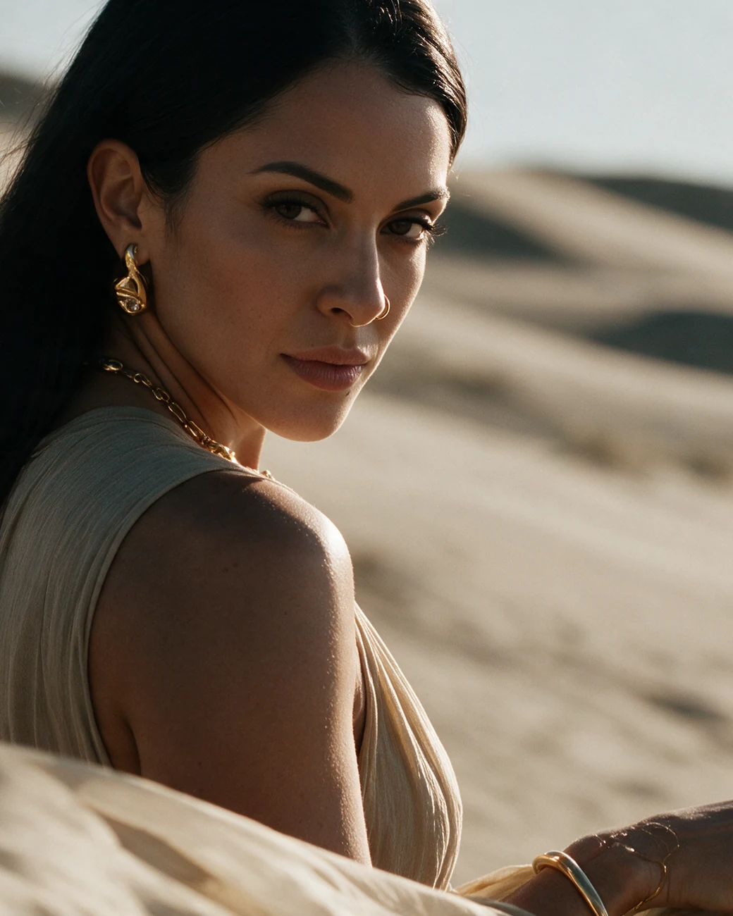 She looks back over a bare shoulder. A gold earring, a necklace and bracelets catch the sun.