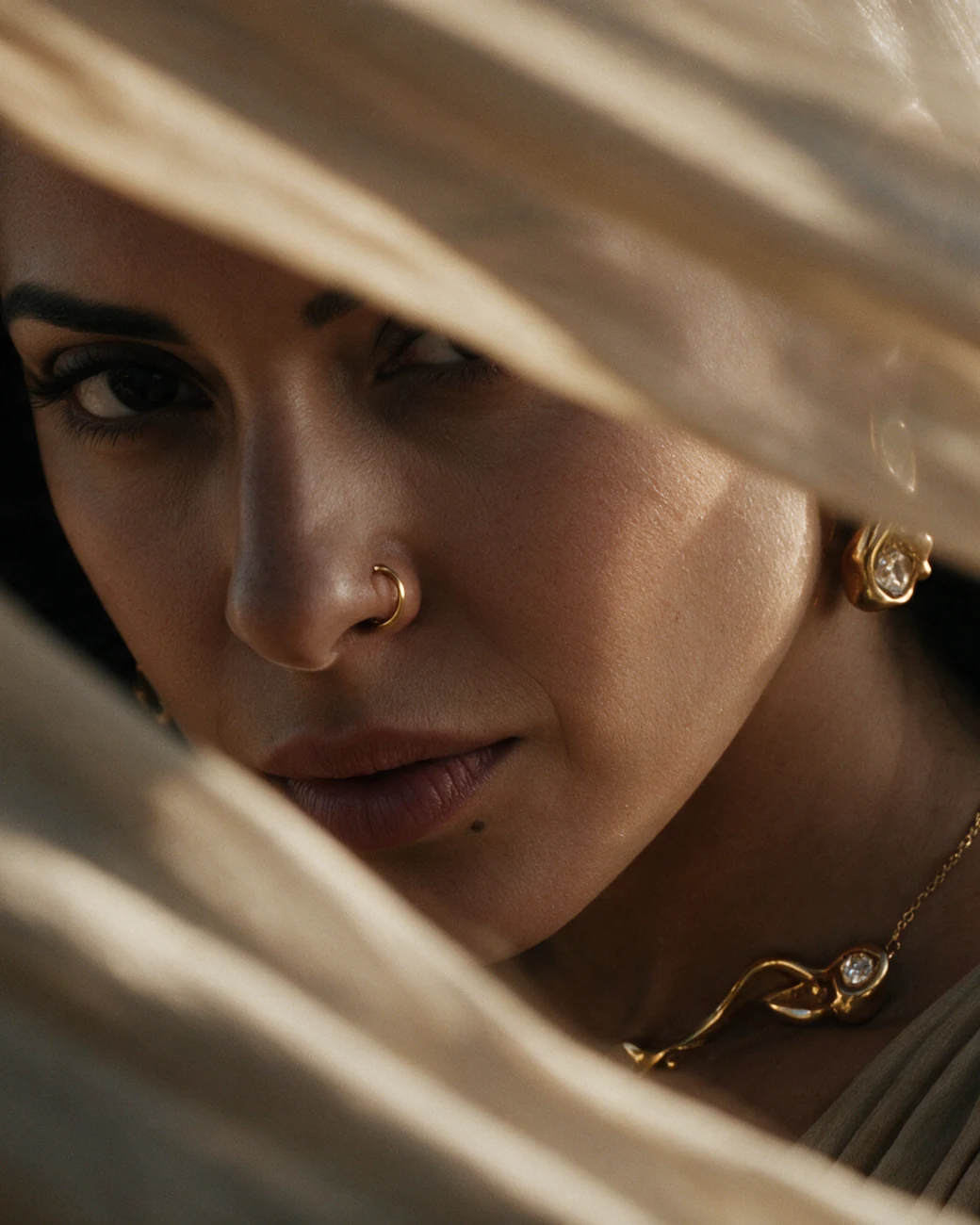 Her face seen through wind-blown fabric, a gold earring and necklace in the light.
